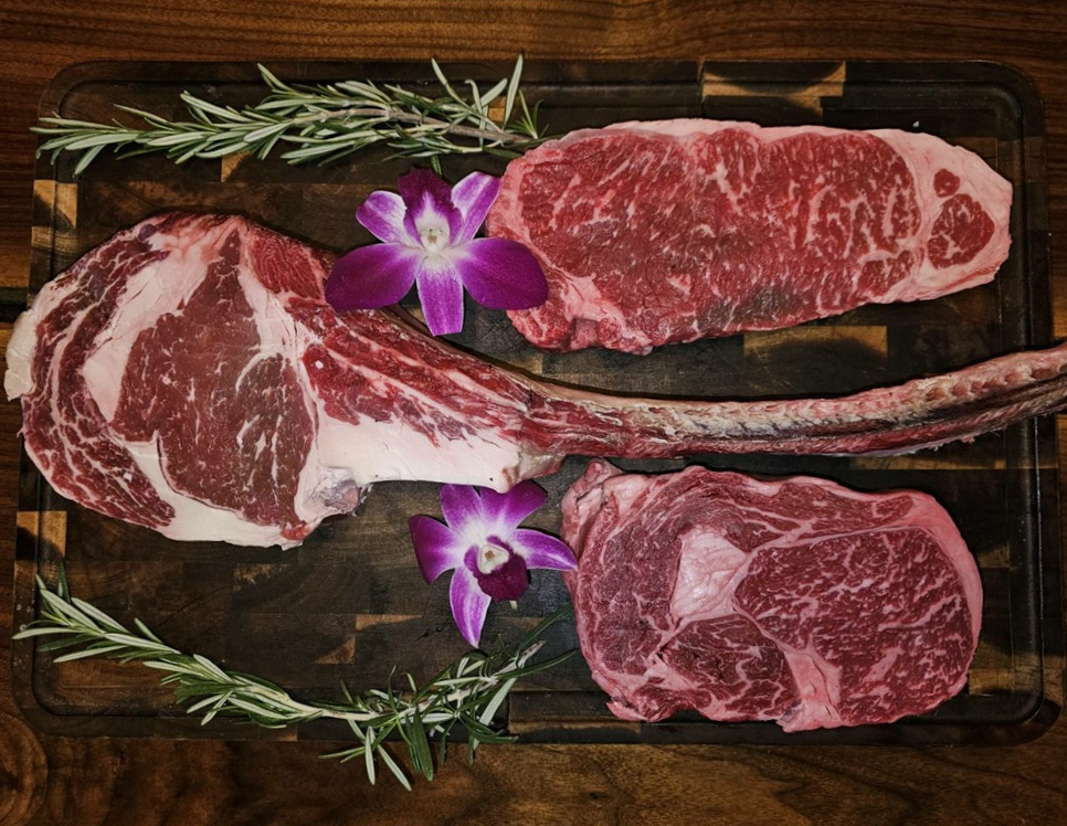 Raw marbled tomahawk, New York strip and ribeye on a butcher block with rosemary and orchids